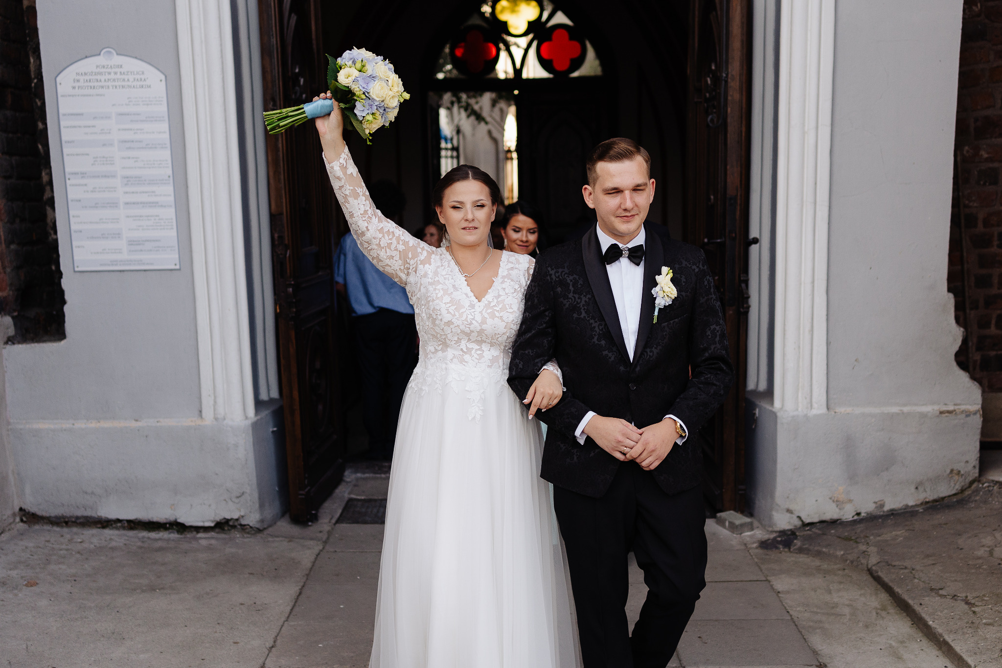 Para młoda opuszcza kościół po ceremonii. Panna młoda trzyma bukiet w górze, obok niej pan młody w eleganckim smokingu. W tle widoczne detale architektoniczne.