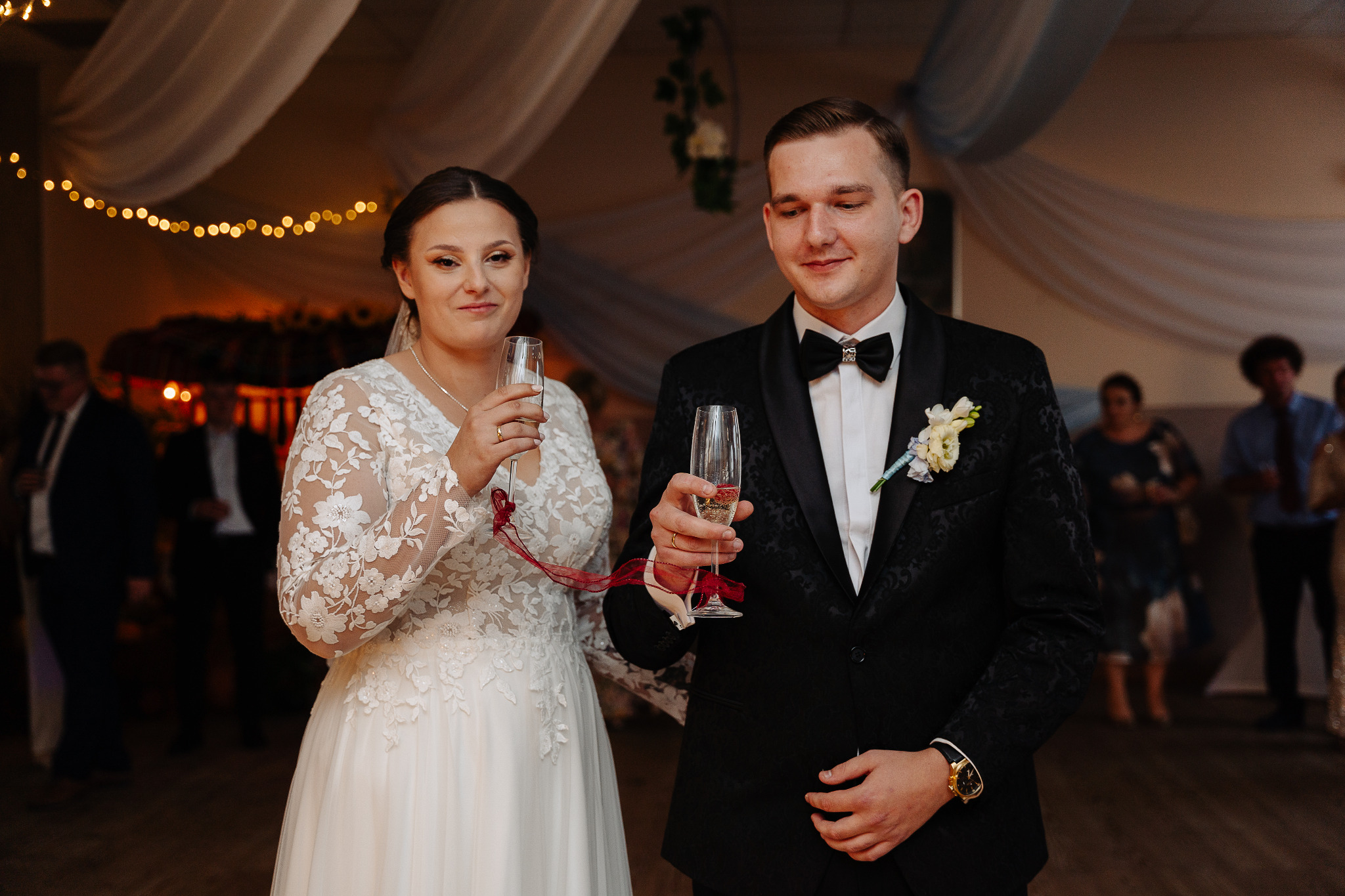 Elegancka para młoda wznosi toast szampanem podczas wesela. Panna młoda w koronkowej sukni, pan młody w smokingu z muchą. Dekoracje i goście w tle.