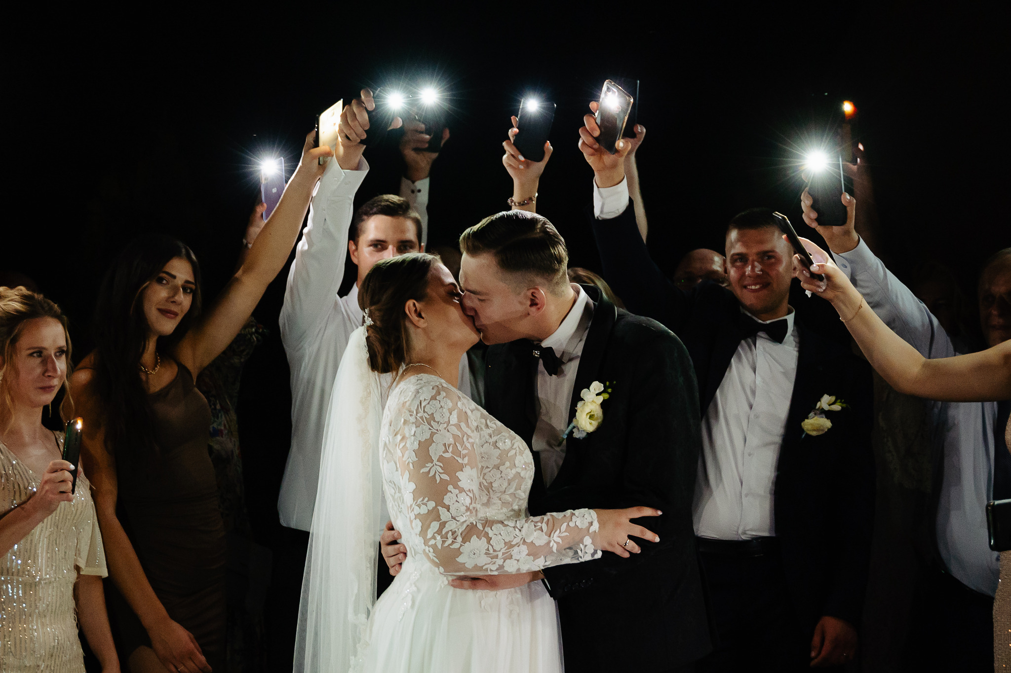 Pocałunek Młodej Pary w otoczeniu gości z telefonami, oświetlającymi ich. Panna Młoda w koronkowej sukni, Pan Młody w smokingu z butonierką.