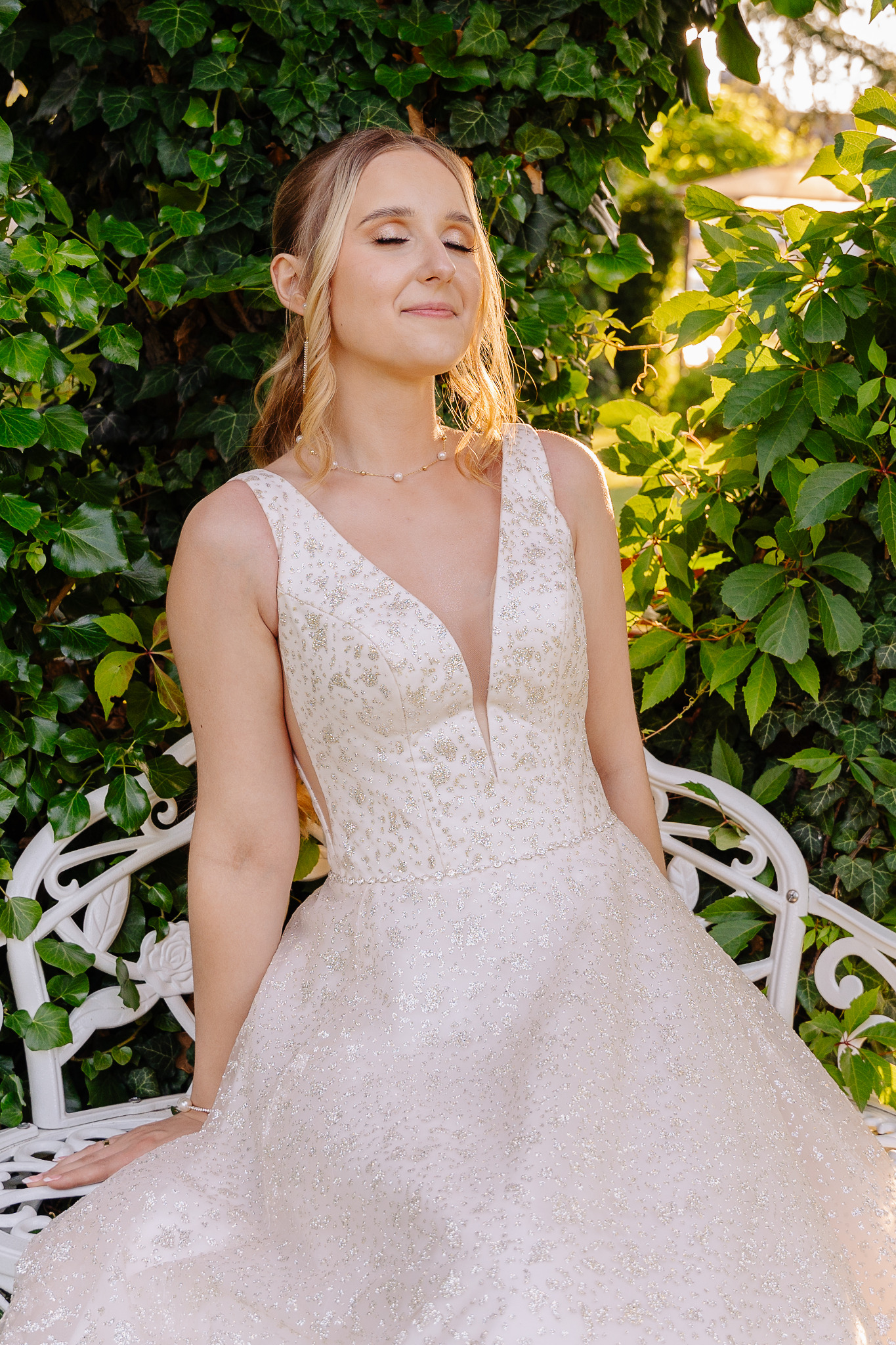 Panna młoda w sukni ślubnej z głębokim dekoltem, siedząca na białej ławce w ogrodzie, z zamkniętymi oczami i uśmiechem, w promieniach słońca.
