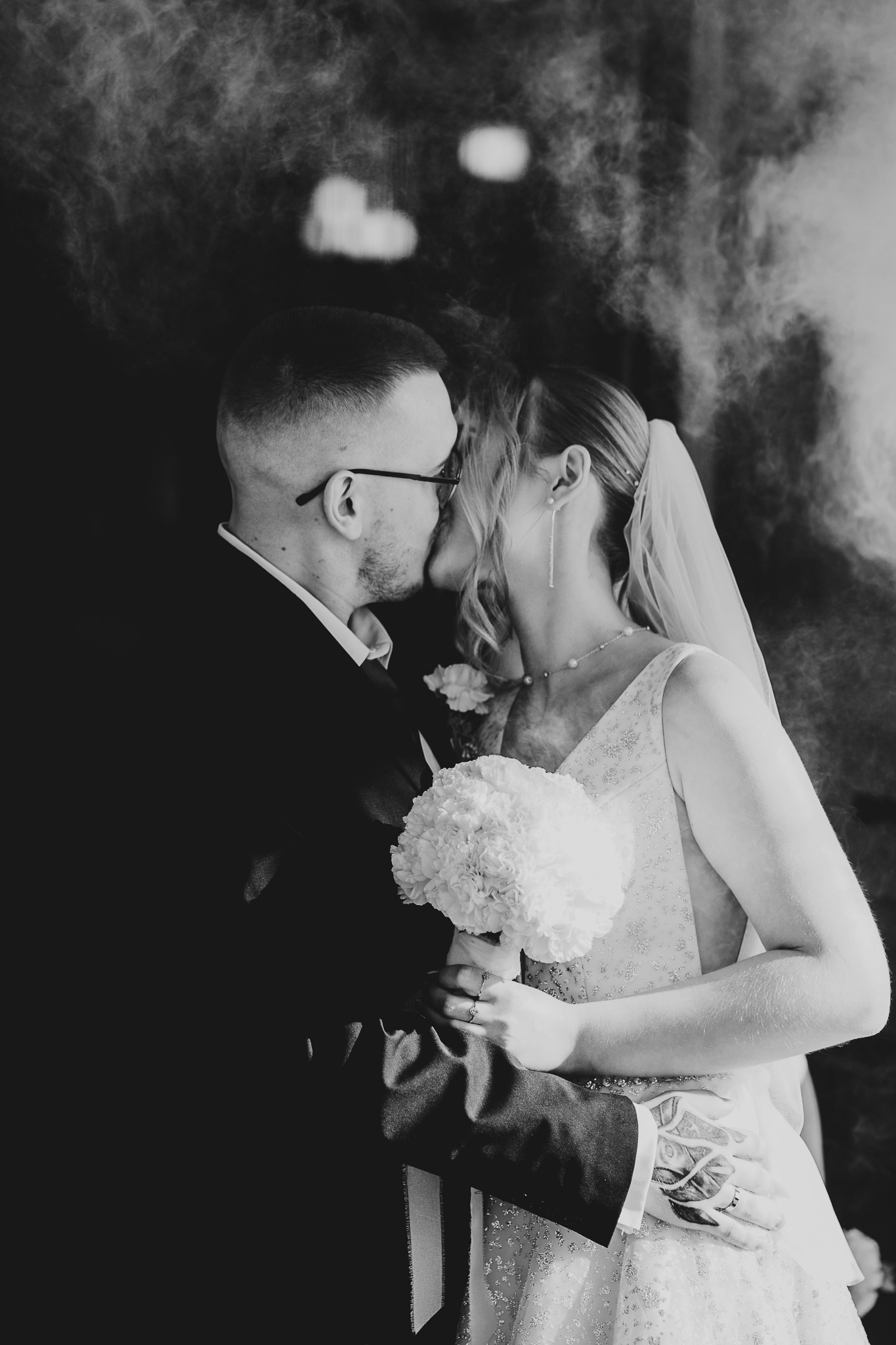 Czarno-białe zbliżenie całującej się pary młodej. Panna młoda trzyma bukiet białych kwiatów. W tle delikatne rozmycie, nadające scenie romantyczny charakter.