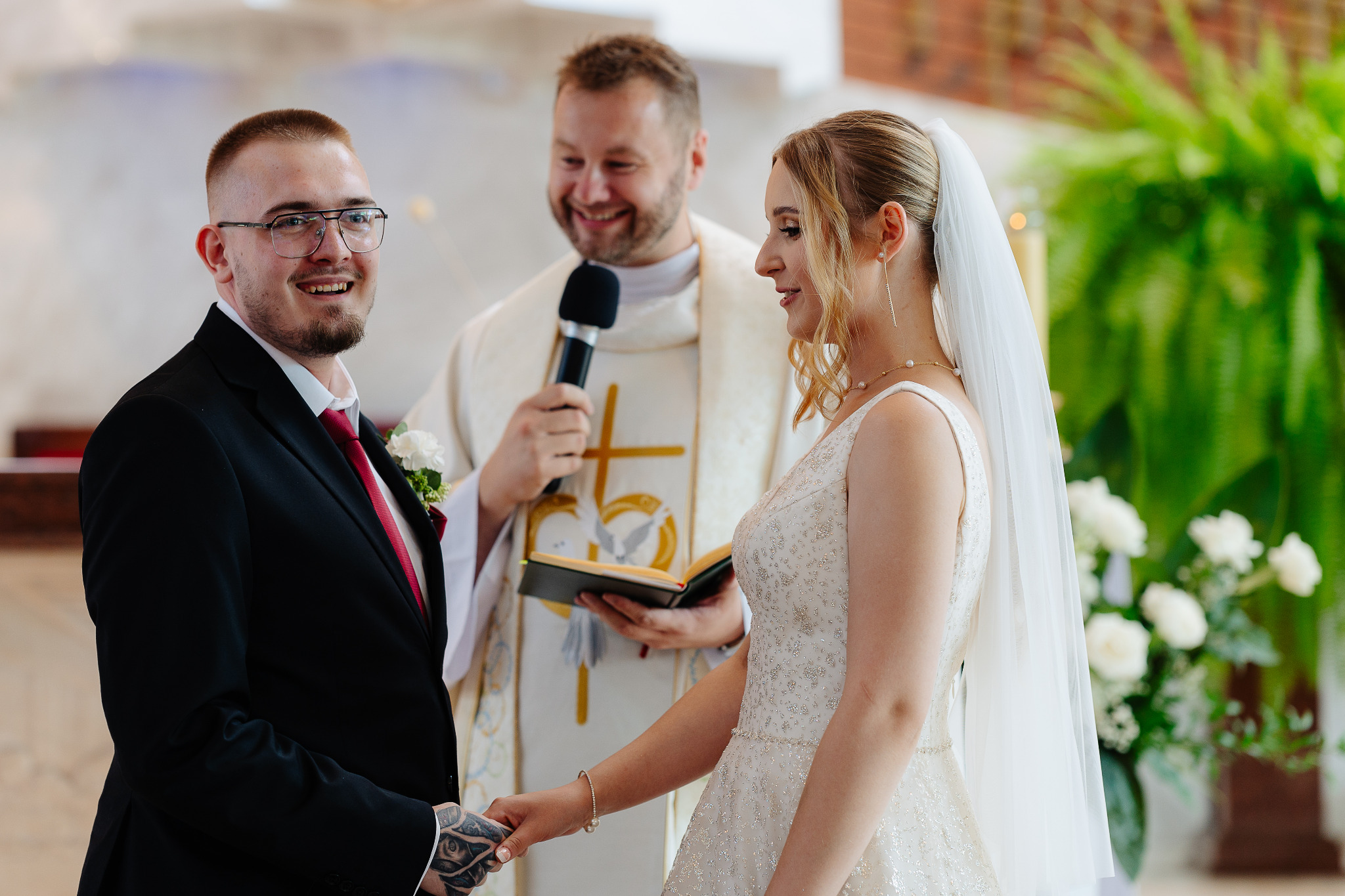 Para młoda trzymająca się za ręce podczas ceremonii ślubnej w kościele, z uśmiechniętym księdzem w tle, z widocznym tatuażem na dłoni pana młodego.
