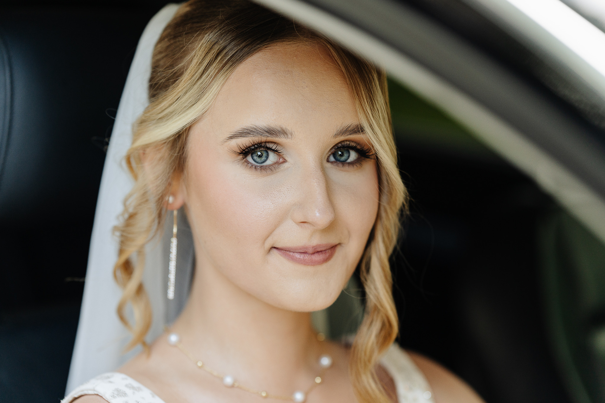 Portret panny młodej w samochodzie, z delikatnym makijażem, blond włosami i welonem. Widoczne kolczyki i naszyjnik z pereł. Uśmiech na twarzy.