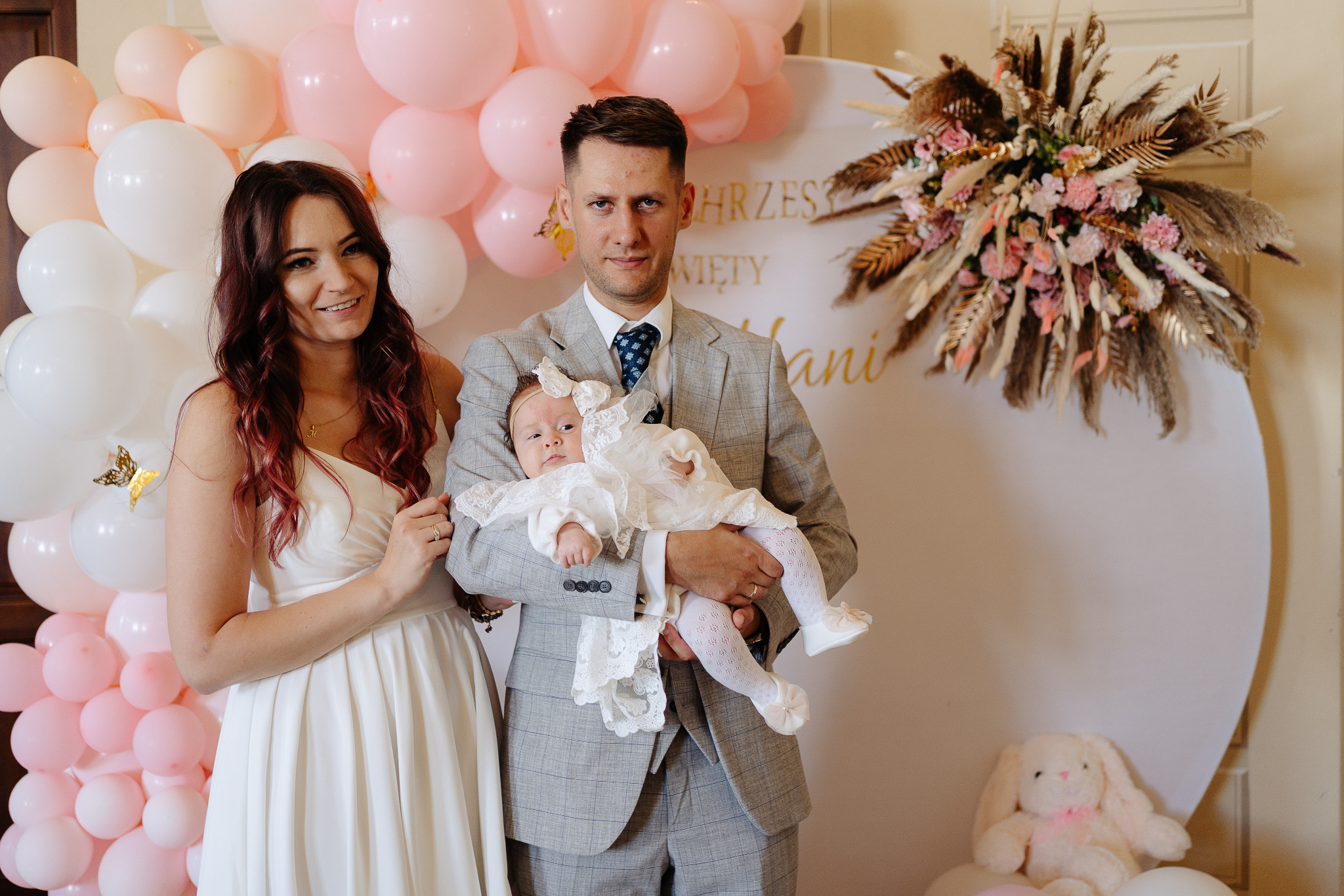 Rodzinne zdjęcie z uroczystości chrztu: rodzice z niemowlęciem na tle dekoracji z balonów i kwiatów w stylu boho. Ojciec w szarym garniturze, matka w białej sukience.