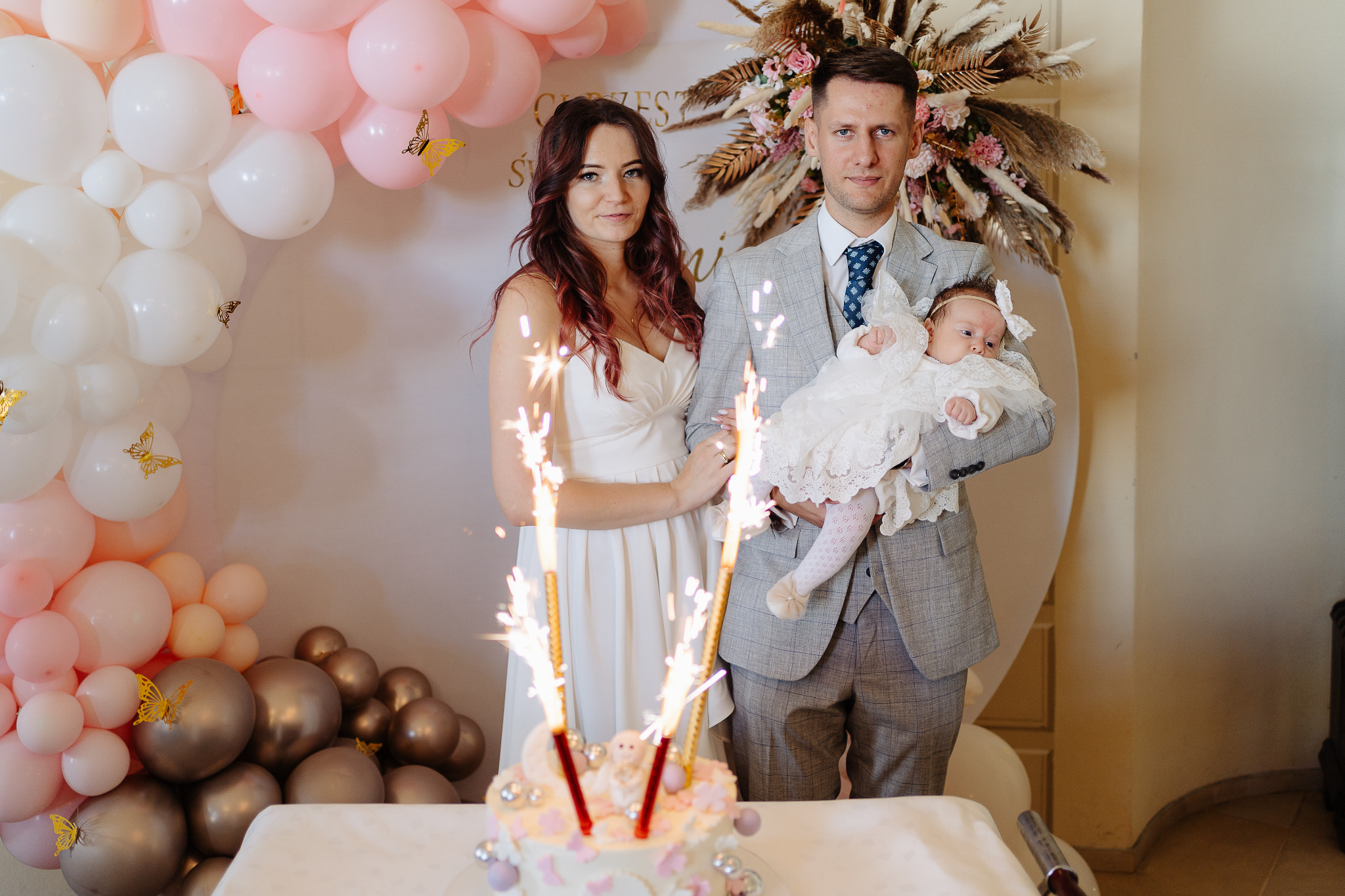 Rodzinne zdjęcie z uroczystości chrztu: rodzice z dzieckiem przy torcie ze świecami, dekoracje z balonów w odcieniach bieli, różu i złota.