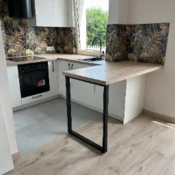 Nowoczesna kuchnia z białymi szafkami, blatem w kolorze jasnego drewna, czarnymi detalami i dekoracyjnym panelem nad blatem z motywem roślinnym. Dodatkowy blat wsparty na metalowej konstrukcji...