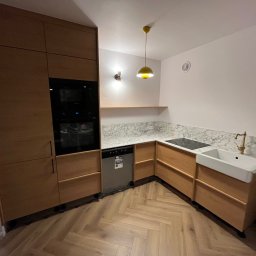 Michał Dominiak Usługi stolarskie - Jasna kuchnia z drewnianymi szafkami, marmurowym blatem i żółtą lampą. Uwagę przykuwa biała, ceramiczna umywalka w stylu retro i parkiet ułożony w jodełkę.