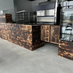 Bar w restauracji z drewnianym frontem o opalonym wyglądzie, widoczne metalowe półki i przeszklona witryna chłodnicza.