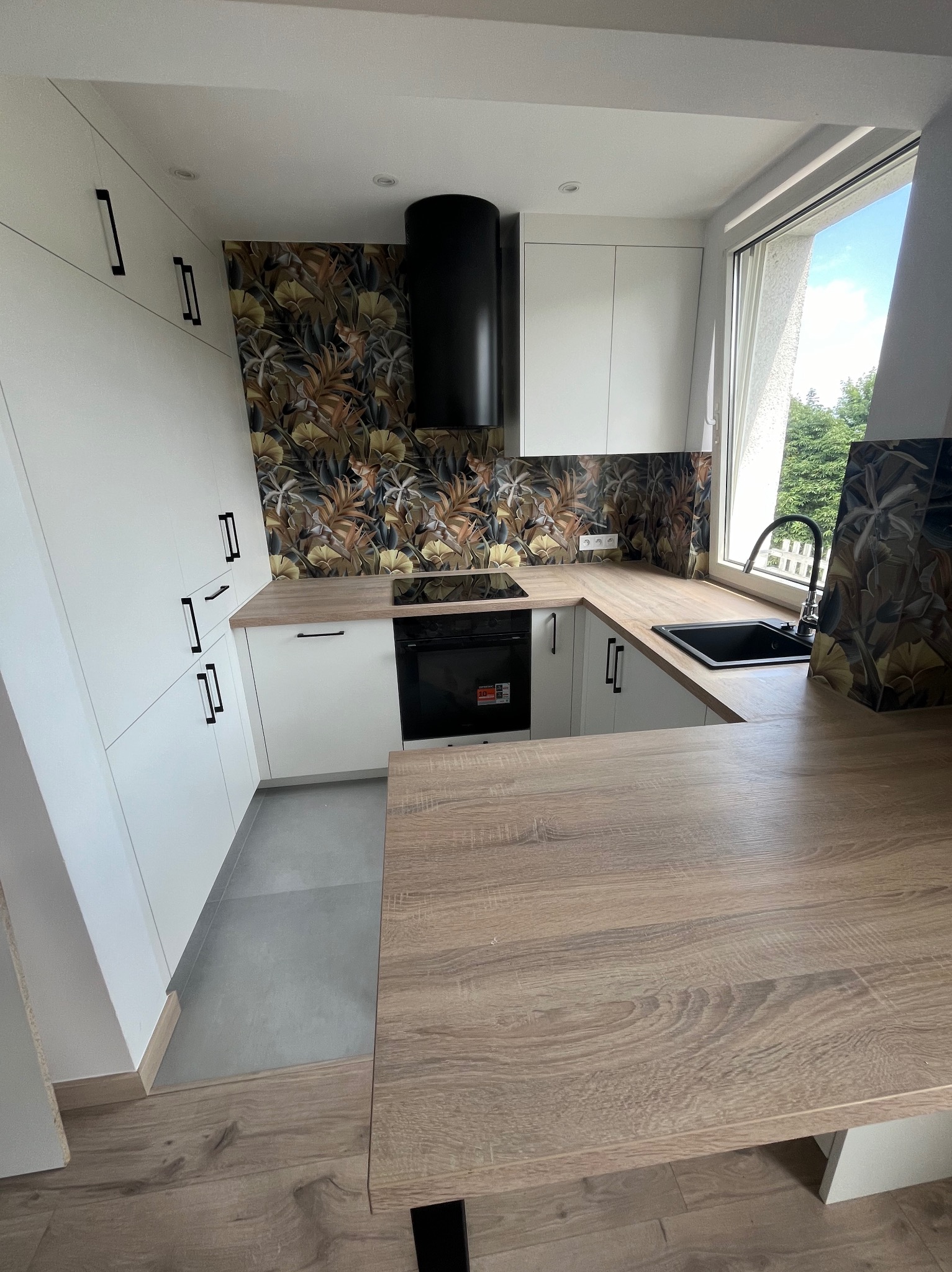 Nowoczesna kuchnia w zabudowie z białymi szafkami, czarnymi uchwytami i blatem imitującym drewno, wykończona tapetą z motywem roślinnym i czarnym okapem.