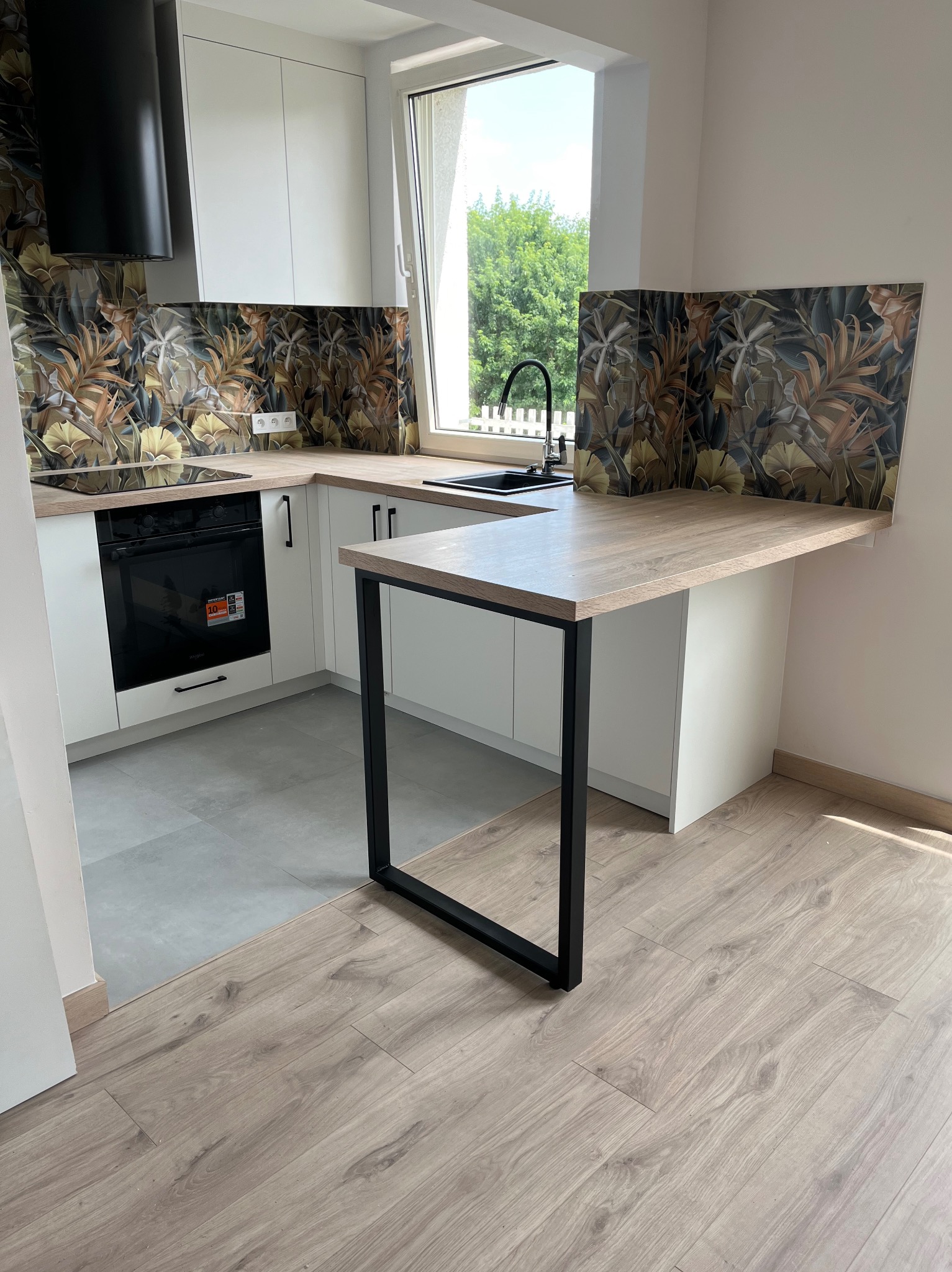 Nowoczesna kuchnia z białymi szafkami, blatem w kolorze jasnego drewna, czarnymi detalami i dekoracyjnym panelem nad blatem z motywem roślinnym. Dodatkowy blat wsparty na metalowej konstrukcji...