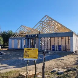 Budowa domu jednorodzinnego w trakcie realizacji: wzniesione ściany z bloczków, konstrukcja drewniana dachu z widocznymi elementami mocującymi, tablica informacyjna budowy na pierwszym planie.