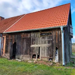 Stara, drewniana stodoła z nowym, czerwonym dachem z dachówki ceramicznej i orynnowaniem, widoczne uszkodzenia desek i częściowe zabicie deskami otworów