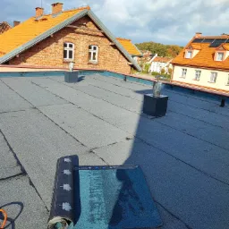 Rolka niebieskiej papy na szarym, płaskim dachu z kominkami wentylacyjnymi w słoneczny dzień, w tle budynki mieszkalne z pomarańczowymi dachówkami.