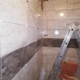 Układanie płytek w łazience, widoczne płytki w odcieniach beżu i brązu z krzyżykami dystansowymi, metalowa drabina oparta o ścianę, częściowo zakryta toaleta.