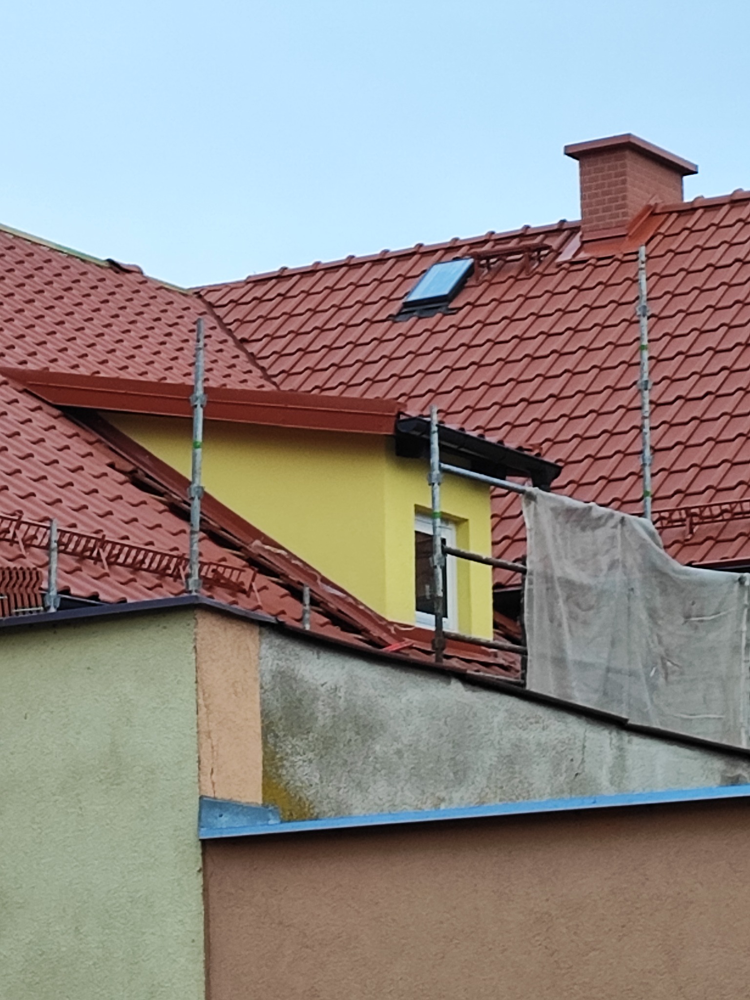 Częściowy widok dachu z czerwoną dachówką, oknem dachowym i kominem, fragment żółtej ściany szczytowej z oknem, rusztowanie i plandeka zabezpieczająca.