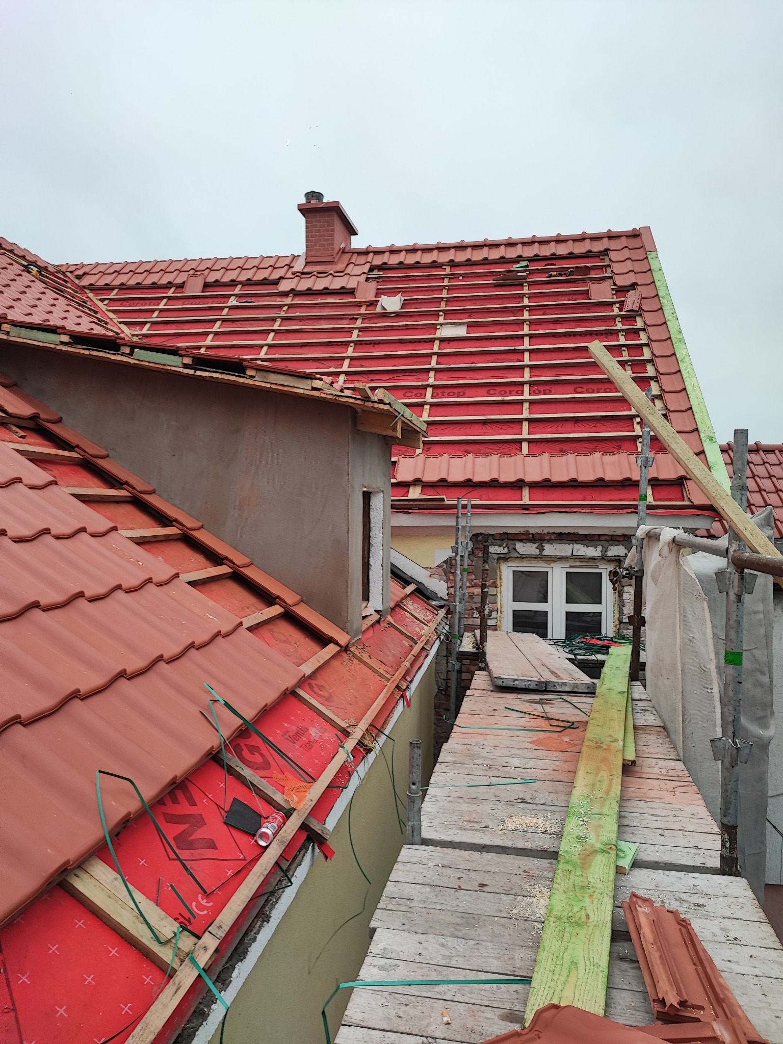 Czerwony dach w trakcie krycia z widocznymi łatami, membraną i częściowo ułożoną dachówką, rusztowanie z desek wzdłuż ściany, komin w tle.