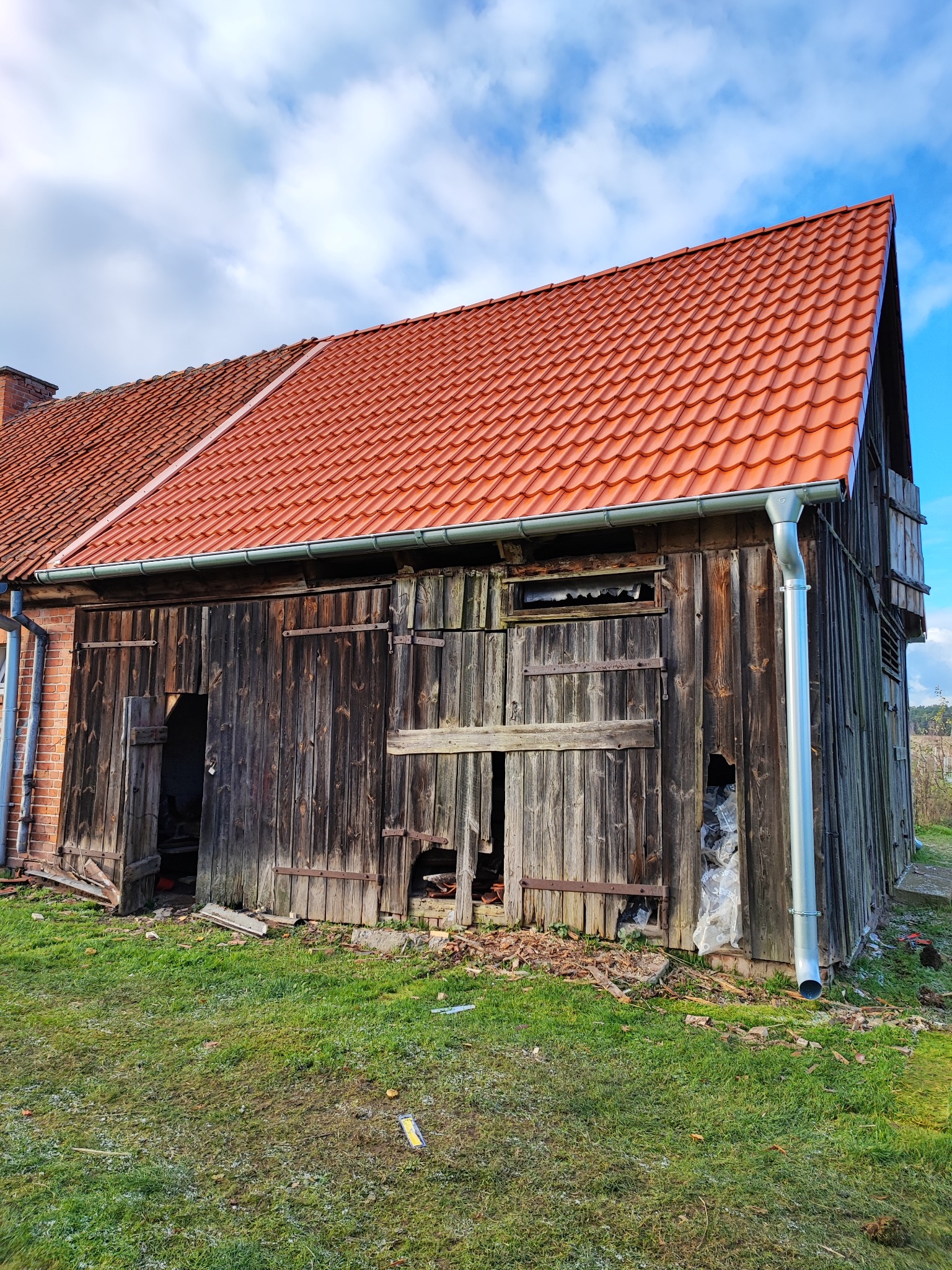 Stara, drewniana stodoła z nowym, czerwonym dachem z dachówki ceramicznej i orynnowaniem, widoczne uszkodzenia desek i częściowe zabicie deskami otworów