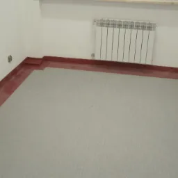 Szara, jednolita powierzchnia podłogi z PCV, otoczona czerwoną listwą przypodłogową w pokoju z białymi ścianami i grzejnikiem.