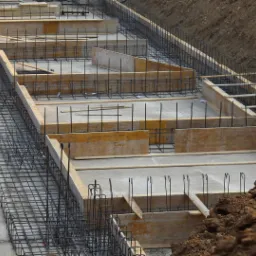 Wykop fundamentowy z deskowaniem i zbrojeniem prętami stalowymi, widok z góry na plac budowy.