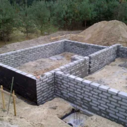 Wznoszenie ścian fundamentowych z bloczków betonowych na budowie domu jednorodzinnego, widoczna czarna izolacja pionowa na fragmencie ściany oraz wystające pręty zbrojeniowe.