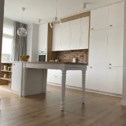 Jasna kuchnia z białymi frontami szafek, drewnianym blatem i ceglaną ścianą nad blatem, oświetlona wiszącymi lampami kulami, z drewnianą podłogą i wyspą z blatem na wysokich nogach.