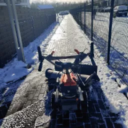 Szczotka mechaniczna do usuwania śniegu na chodniku z kostki brukowej, częściowo odśnieżonym, w słoneczny dzień przy metalowym ogrodzeniu.