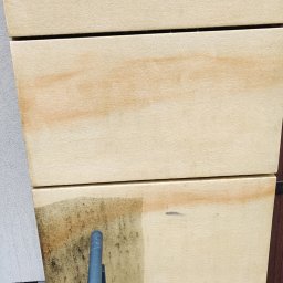 DreamClean Sp. z o.o. - Częściowe oczyszczenie elewacji z jasnych płyt, widoczne różnice w kolorze i zabrudzeniu przed i po czyszczeniu, z fragmentem narzędzia czyszczącego w dolnej części kadru.