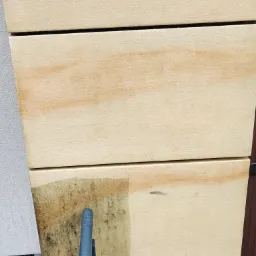 Częściowe oczyszczenie elewacji z jasnych płyt, widoczne różnice w kolorze i zabrudzeniu przed i po czyszczeniu, z fragmentem narzędzia czyszczącego w dolnej części kadru.