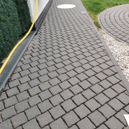 DreamClean Sp. z o.o. - Ułożona kostka brukowa w kolorze grafitowym, z widocznym obrzeżem, trawą i fragmentem węża ogrodowego zwiniętego na ścianie budynku.