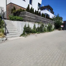 Nowoczesny budynek z drewnianą elewacją i schodami wejściowymi, otoczony zielenią i drewnianym ogrodzeniem, widok z chodnika wyłożonego kostką brukową.