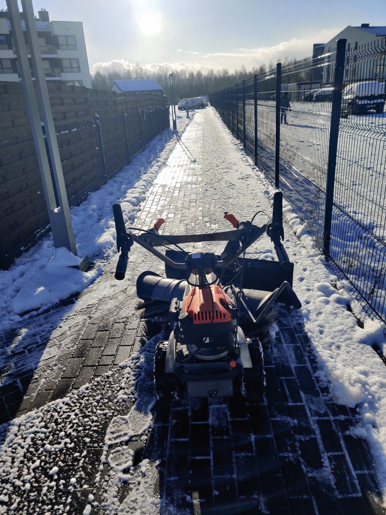Szczotka mechaniczna do usuwania śniegu na chodniku z kostki brukowej, częściowo odśnieżonym, w słoneczny dzień przy metalowym ogrodzeniu.