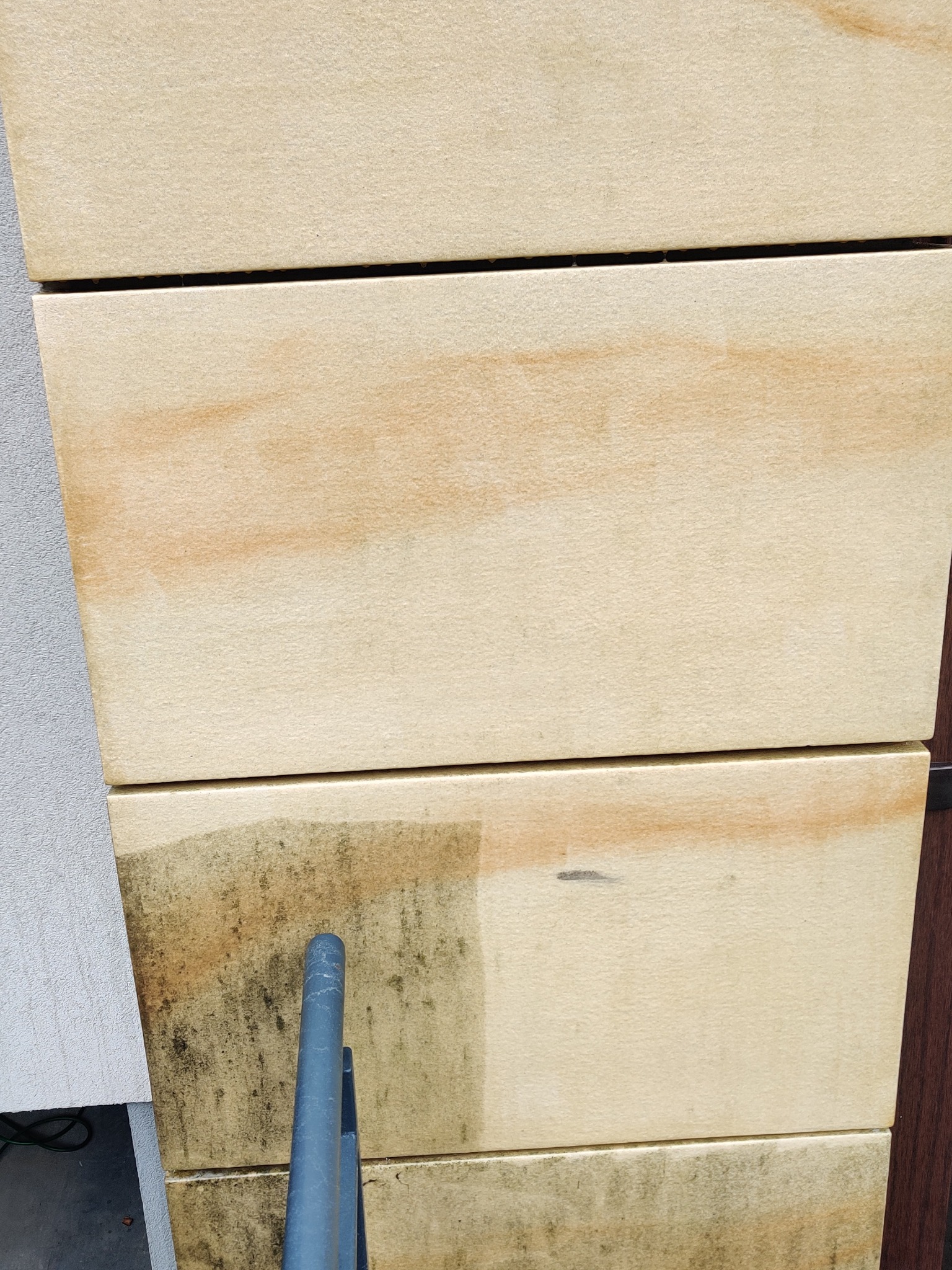 Częściowe oczyszczenie elewacji z jasnych płyt, widoczne różnice w kolorze i zabrudzeniu przed i po czyszczeniu, z fragmentem narzędzia czyszczącego w dolnej części kadru.
