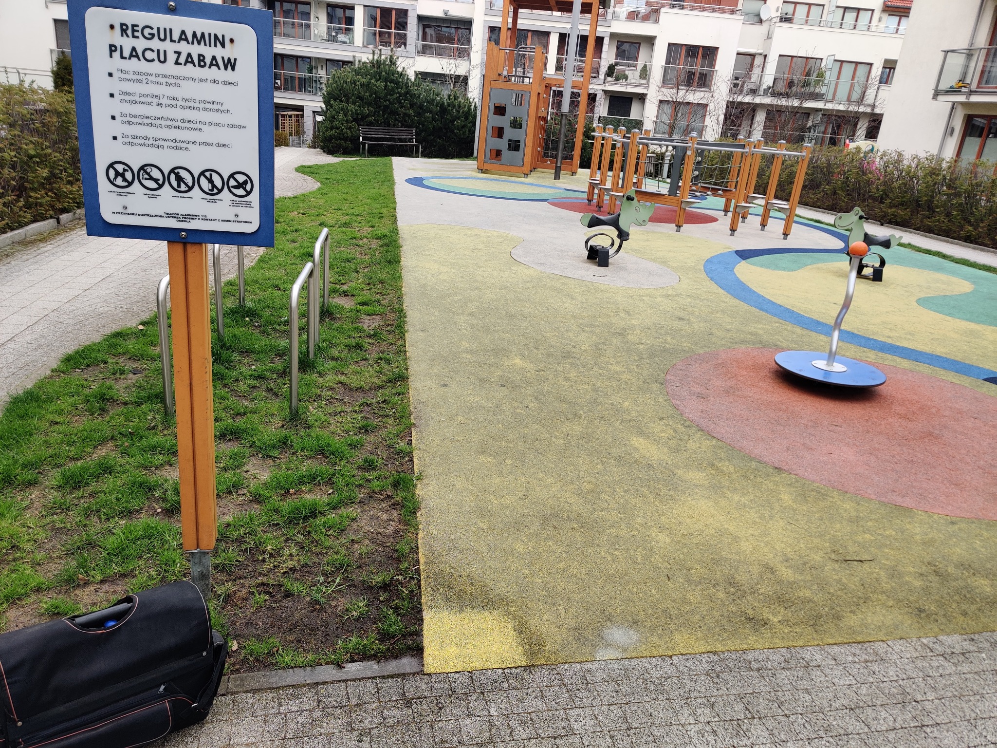 Regulamin placu zabaw na tablicy informacyjnej, obok stojak na rowery i czarna torba podróżna, w tle kolorowa nawierzchnia i urządzenia placu zabaw oraz budynki mieszkalne.
