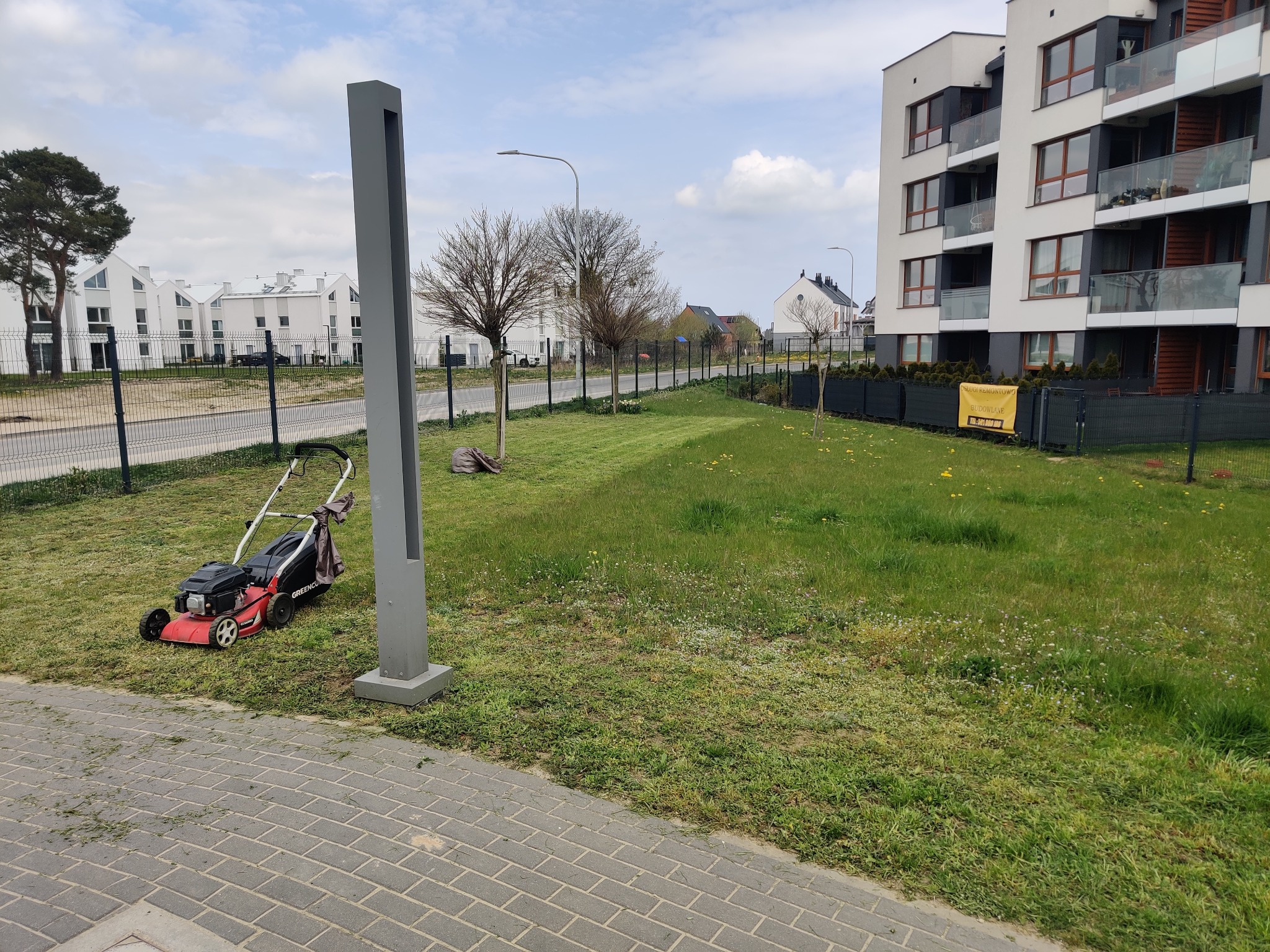 Czerwona kosiarka spalinowa stojąca na trawniku po skoszeniu, widoczne ślady cięcia, w tle budynki mieszkalne i drzewa.