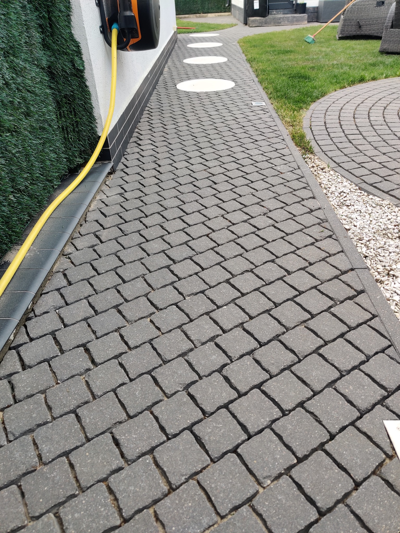 Ułożona kostka brukowa w kolorze grafitowym, z widocznym obrzeżem, trawą i fragmentem węża ogrodowego zwiniętego na ścianie budynku.
