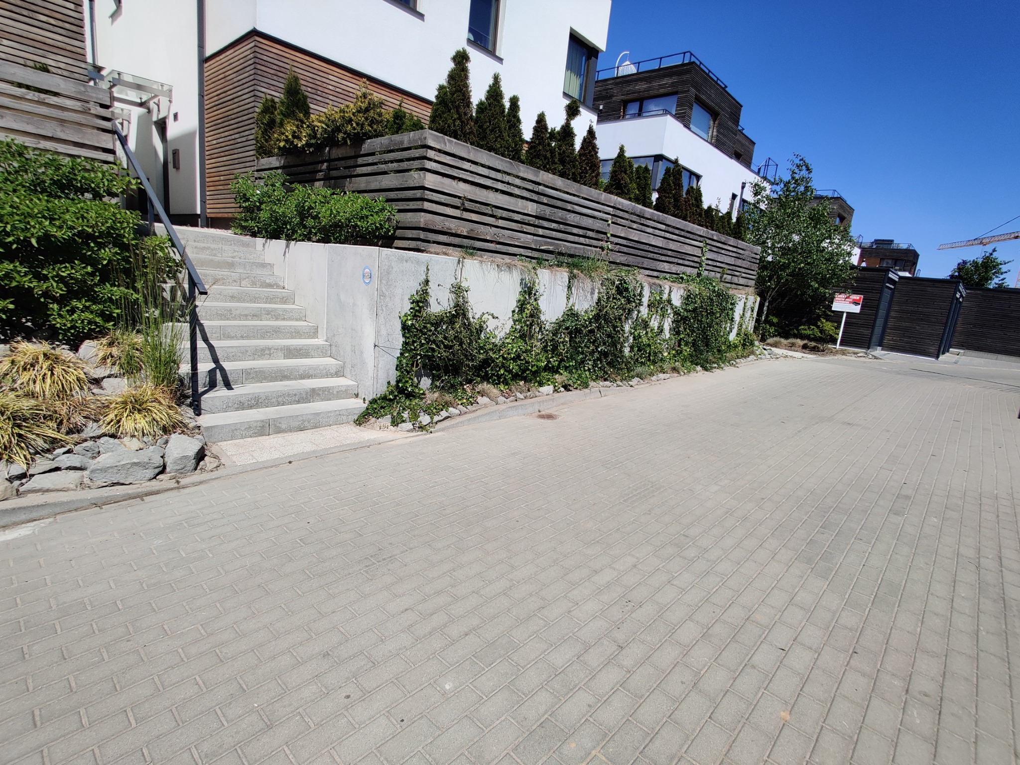 Nowoczesny budynek z drewnianą elewacją i schodami wejściowymi, otoczony zielenią i drewnianym ogrodzeniem, widok z chodnika wyłożonego kostką brukową.