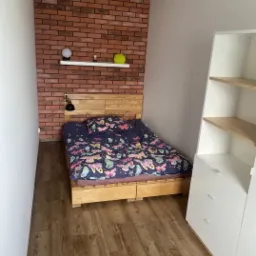 Sypialnia w Częstochowie z łóżkiem na drewnianej ramie, ceglaną ścianą z półką dekoracyjną i białą szafą z szufladami.