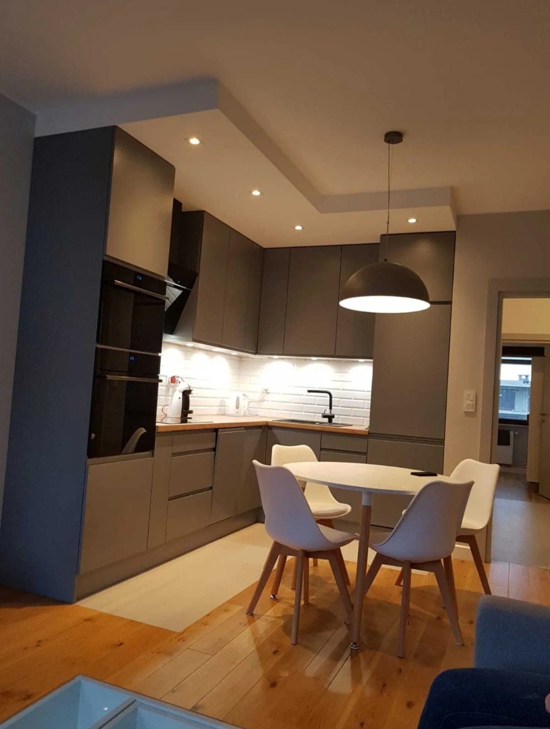 Nowoczesna kuchnia w odcieniach szarości z drewnianą podłogą i białymi krzesłami przy okrągłym stole, oświetlona wiszącą lampą i punktowymi światłami w suficie.