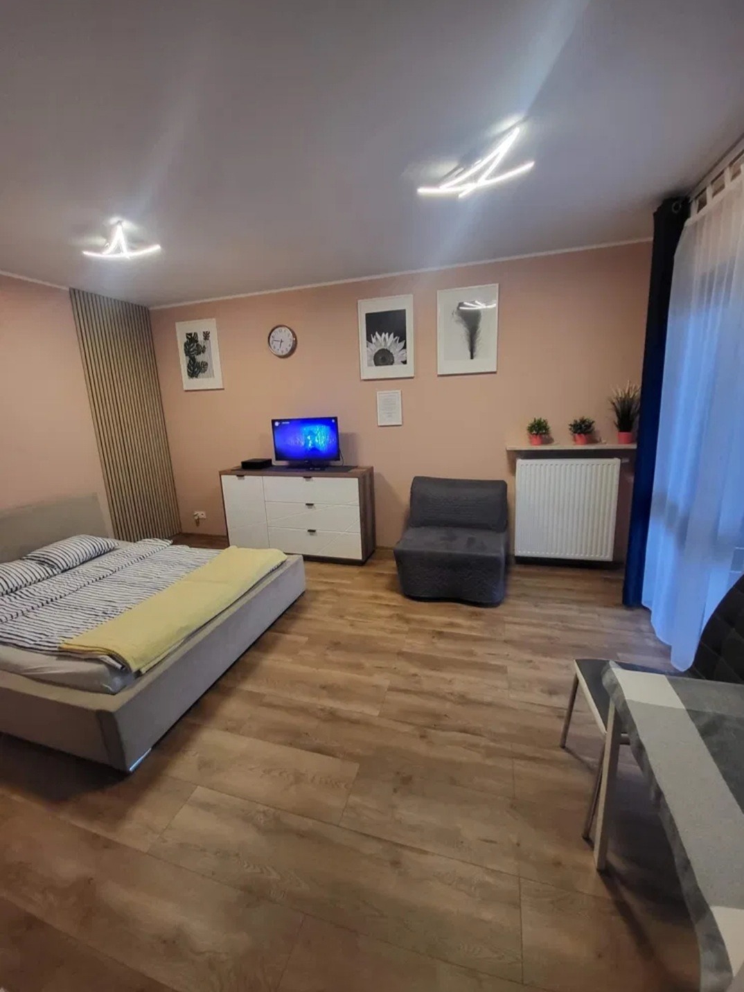 Komfortowo urządzone, jasne studio z drewnianą podłogą, łóżkiem, telewizorem na komodzie i designerskimi lampami sufitowymi.