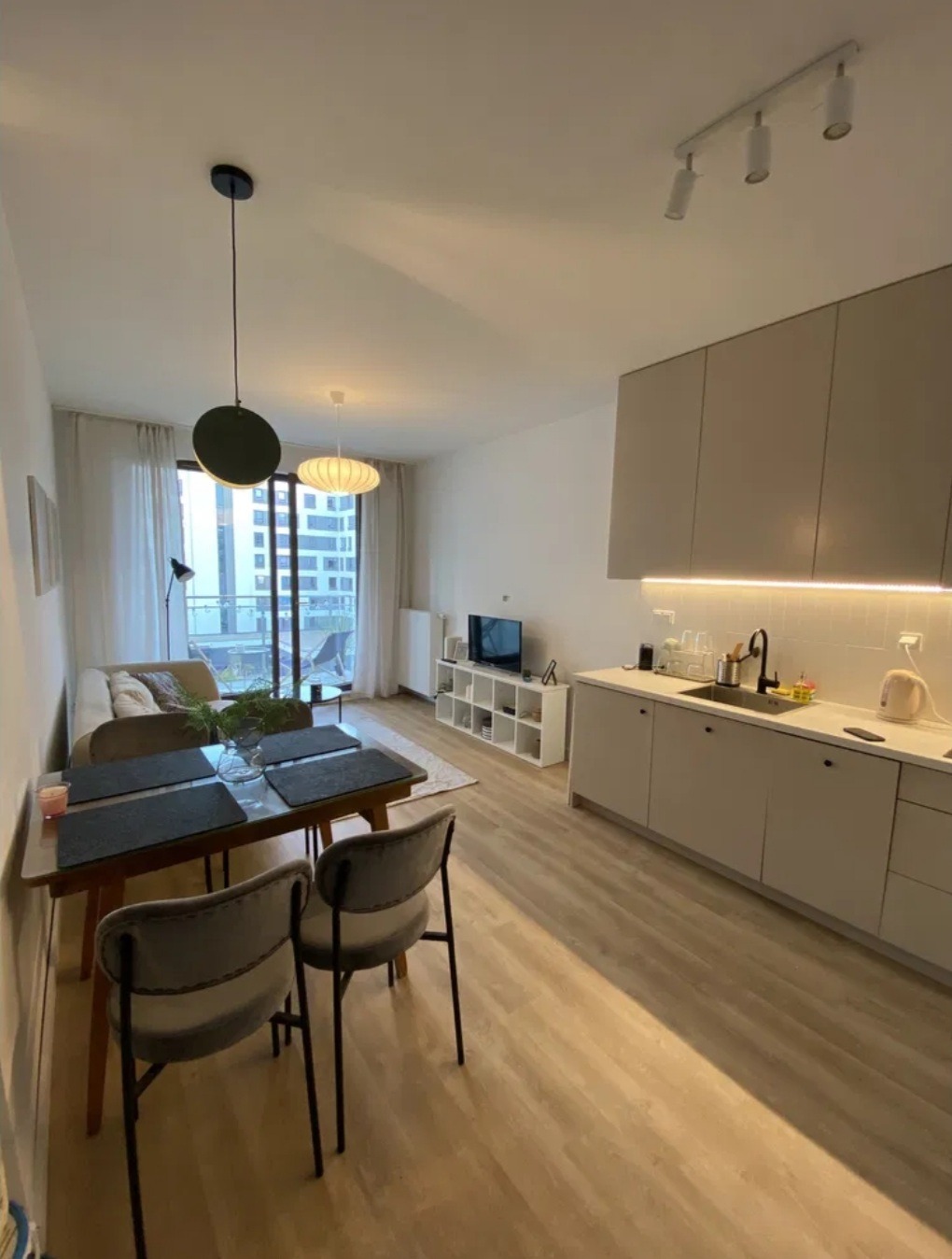 Jasne, nowoczesne wnętrze mieszkania z aneksem kuchennym, drewnianym stołem z szarymi podkładkami i krzesłami, widokiem na balkon i zabudowę miejską za oknem.