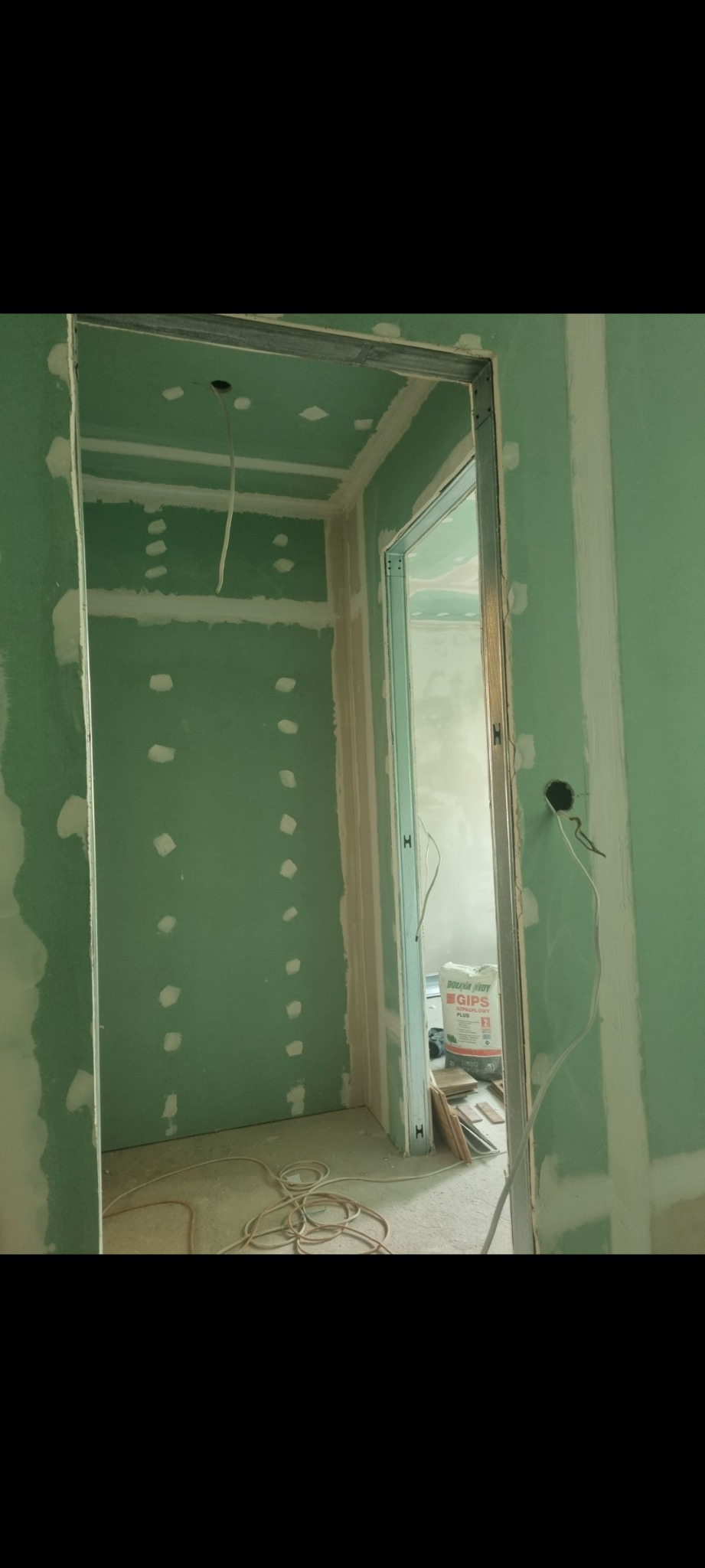 Wnętrze w trakcie montażu płyt kartonowo-gipsowych, widoczne metalowe profile, szpachlowane łączenia i wystające kable elektryczne. Na podłodze leży zwinięty przewód i worek z gipsem.