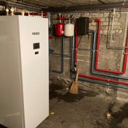 SOLKER sp. z o.o. - Biała pompa ciepła HEIKO w piwnicy z odsłoniętą cegłą, widoczne czerwone i niebieskie rury izolowane, manometry, zbiorniki wyrównawcze oraz stojąca miotła z naturalnego włosia.