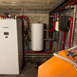 SOLKER sp. z o.o. - Biała pompa ciepła marki Heiko w piwnicy, z widocznymi rurami w izolacji czerwonej i niebieskiej, oraz białym zbiornikiem akumulacyjnym, na tle surowej ściany z cegły i betonowego sufitu.