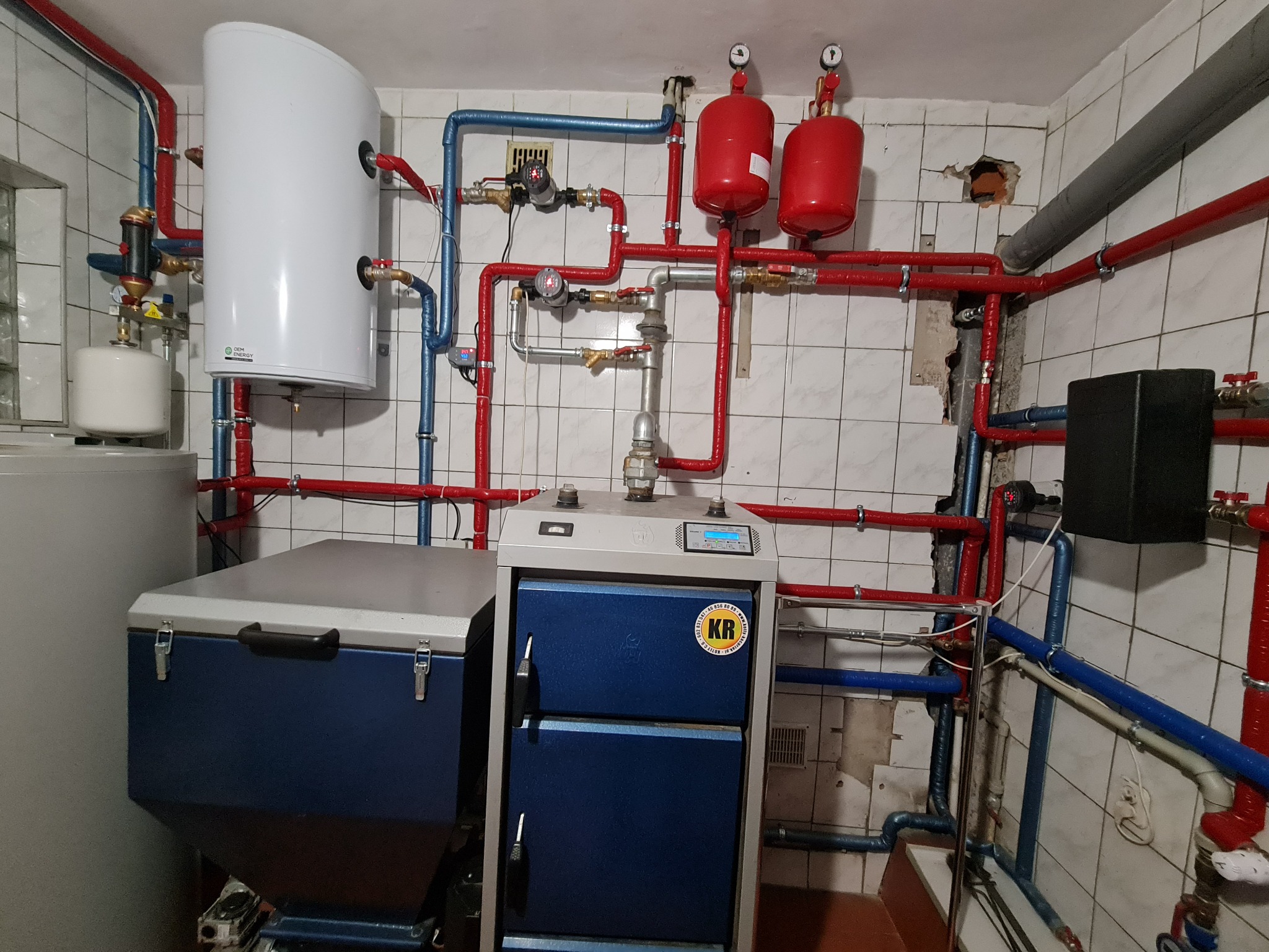 Kotłownia z kotłem stałopalnym i nowym zasobnikiem ciepłej wody, połączone rurami w izolacji czerwonej i niebieskiej, z widocznym wymiennikiem płytowym i dwoma czerwonymi zbiornikami wyrównawczymi.