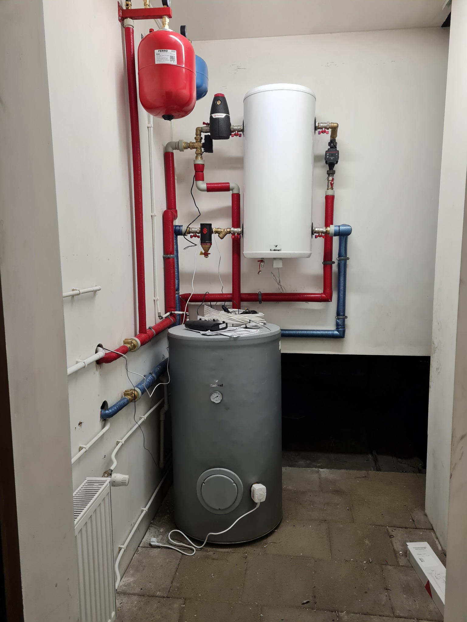 Szary zbiornik akumulacyjny ciepła z podłączonymi rurami, zaworami i białym bojlerem w małym pomieszczeniu z grzejnikiem.