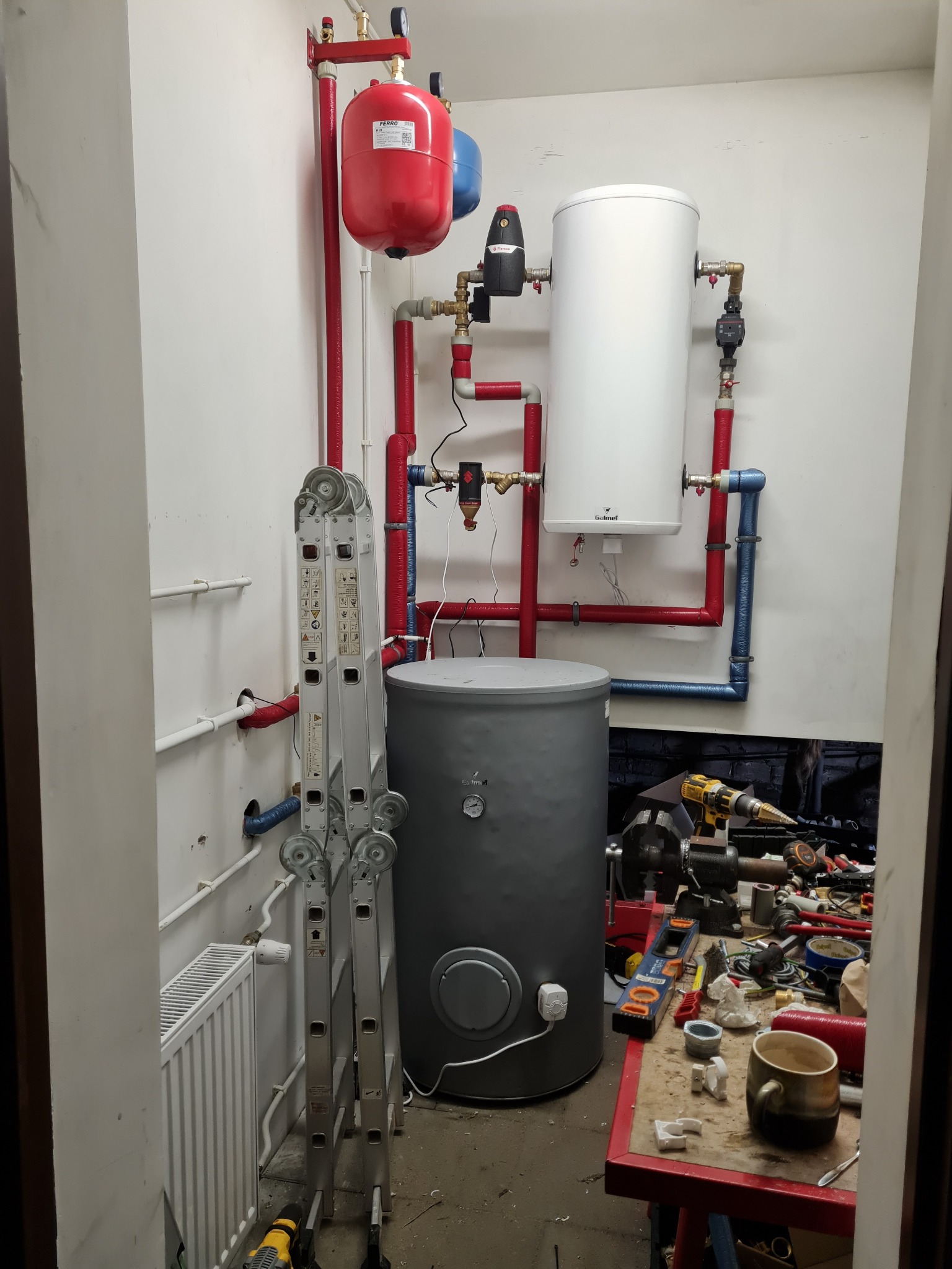Instalacja zbiorników: szary akumulacyjny i biały ciśnieniowy, z czerwonymi i niebieskimi rurami, drabina oparta o ścianę, warsztat z narzędziami w tle.