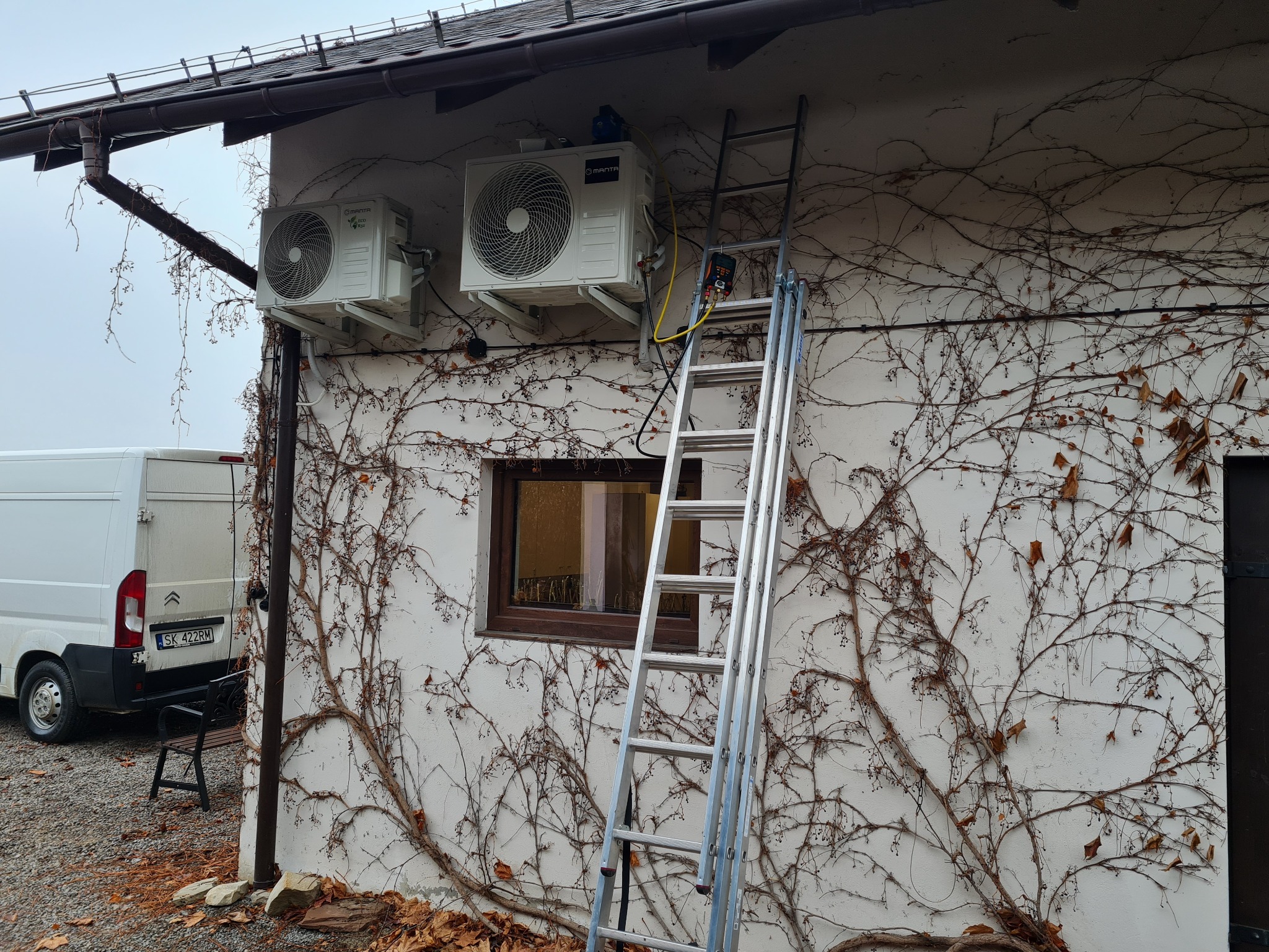 Dwa białe klimatyzatory zamontowane na ścianie budynku porośniętej bluszczem, drabina oparta o ścianę, biały samochód dostawczy w tle.