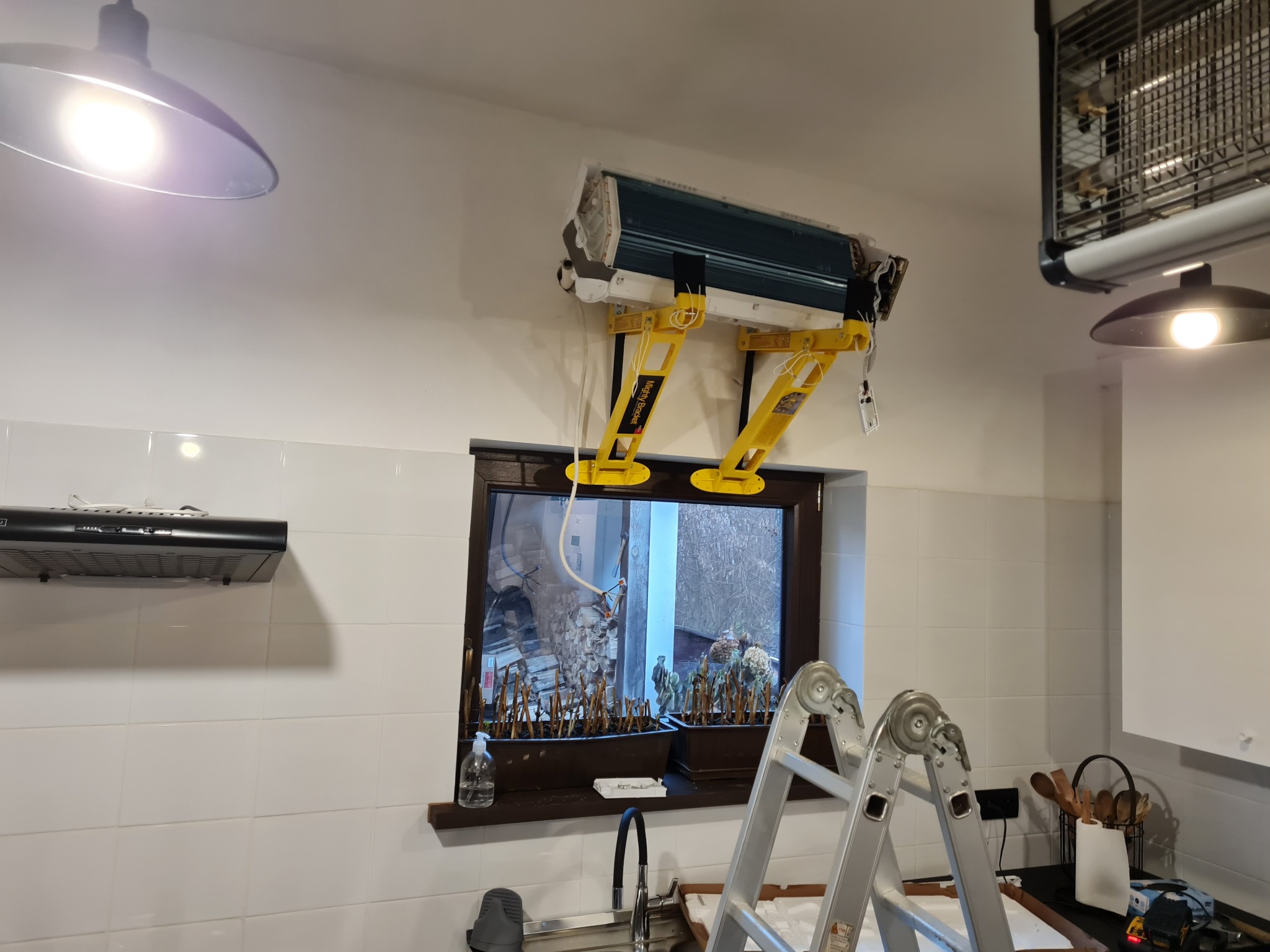Wnętrze kuchni z klimatyzatorem w trakcie montażu na ścianie, podtrzymywanym przez żółte uchwyty montażowe Mighty Bracket, drabina aluminiowa oparta o okno z widokiem na stos drewna.