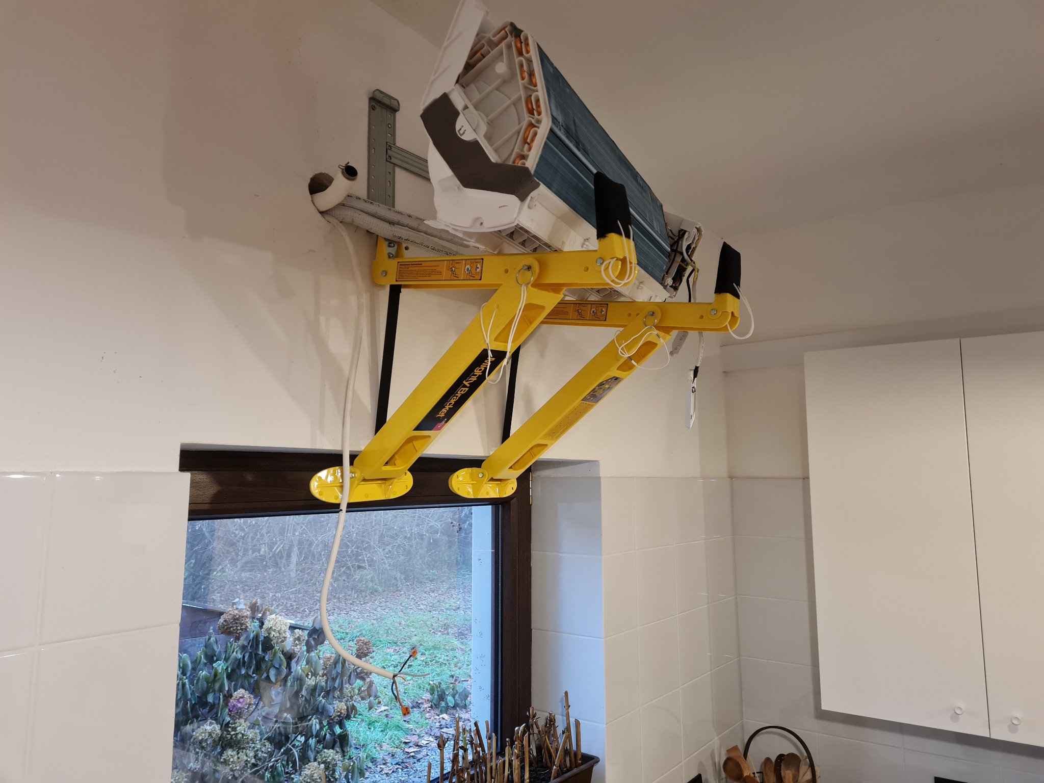 Wnętrze z klimatyzatorem zamontowanym na żółtym podnośniku Mighty Vise Bracket, widoczne instalacje wychodzące przez otwór w ścianie, okno z widokiem na ogród.