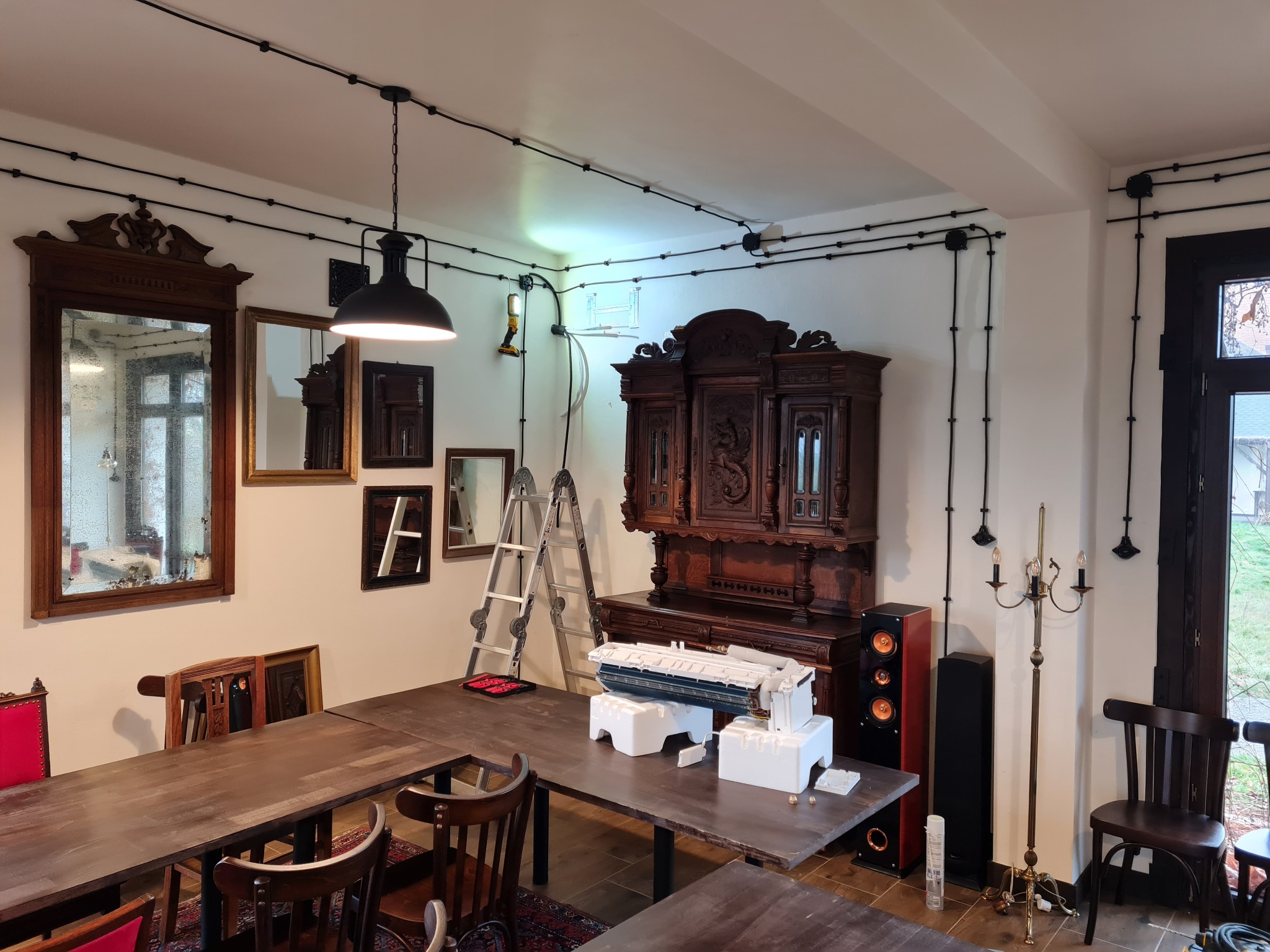 Wnętrze pokoju z antycznymi meblami i rozłożoną jednostką wewnętrzną klimatyzacji na stole, drabina oparta o ścianę, widoczne czarne kable instalacji elektrycznej na suficie i ścianach.